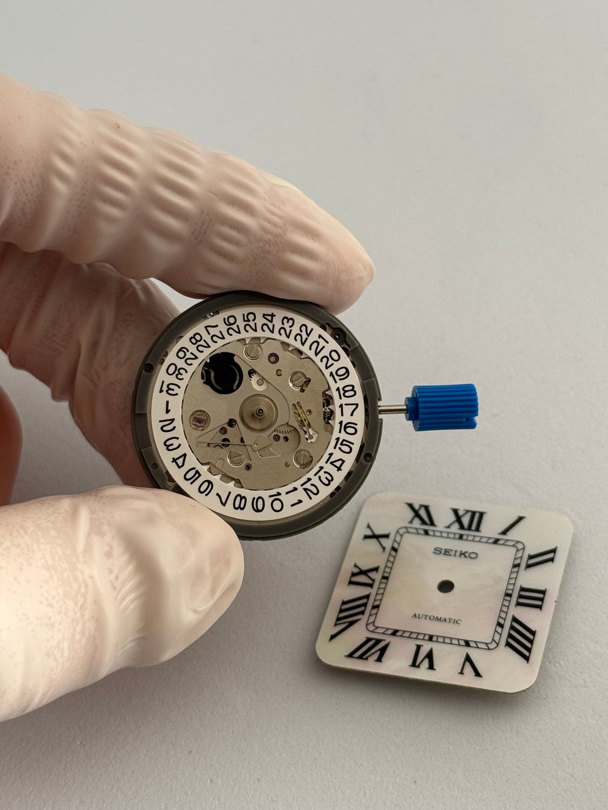 Mouvement automatique Seiko NH35 et son disque de date, tenu entre des doigts gantés, à côté d'un cadran nacré à chiffres romains.